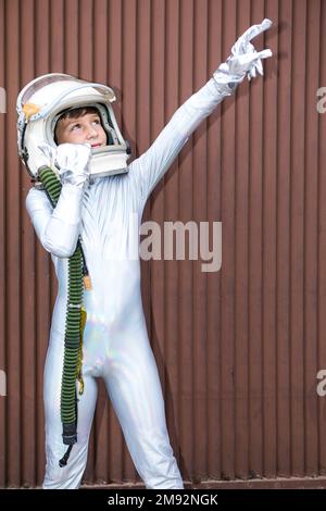 Curieux préadolescent dans combinaison d'espace debout devant le mur côtelé brun et pointant vers le haut Banque D'Images