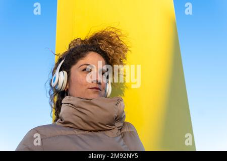 Faible angle de la femme attentionnés en vêtements d'extérieur écoutant votre chanson préférée sur un casque blanc, en regardant loin du ciel bleu sans nuages Banque D'Images