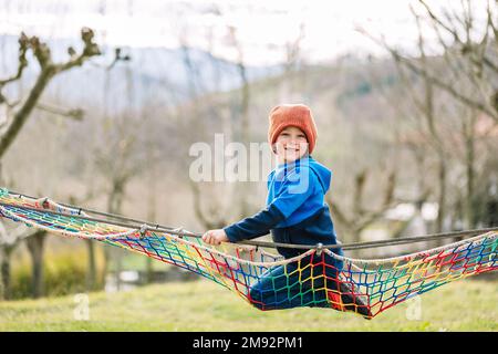 Positif garçon dans des vêtements chauds et chapeau souriant heureux et regardant la caméra tout en rampant sur le filet de corde dans le parc d'automne Banque D'Images
