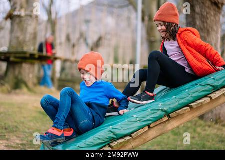 Des enfants gais dans des vêtements chauds souriant et glissant tout en jouant sur l'aire de jeux ensemble pendant le week-end au parc Banque D'Images