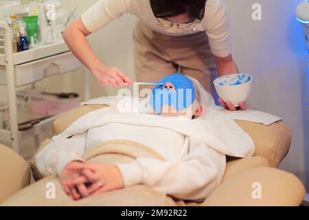De dessus de la femme cosmétologue dans les lunettes application du masque alginate par la spatule sur le visage du client pendant les soins de la peau Banque D'Images