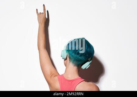 Vue arrière d'une petite femelle non reconnaissable avec des cheveux courts bleus uniques qui écoutent vos chansons préférées sur des écouteurs en studio, montrant des gestu rock and roll Banque D'Images