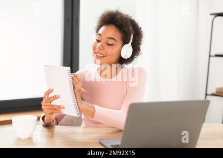 Une jeune femme de course mixte souriante et rêveuse, dans un casque sans fil, prend des notes Banque D'Images
