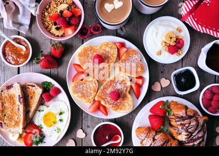 Scène de table pour le brunch de la Saint-Valentin ou de la fête des mères. Vue de dessus sur un fond en bois sombre. Crêpes en forme de cœur, œufs et assortiment de plats sur le thème de l'amour. Banque D'Images