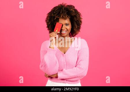 Bonne millénaire afro-américaine curly femme shopaholic dans décontracté met carte de crédit à l'oeil, a l'amusement Banque D'Images