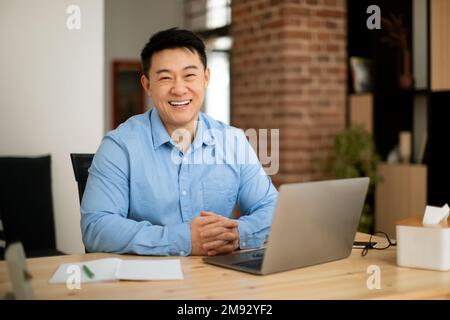 Portrait d'un homme asiatique mûr heureux avec un ordinateur portable, assis à une table dans le salon et souriant à l'appareil photo, espace libre Banque D'Images