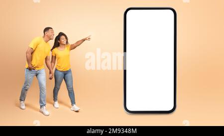 Une offre géniale. Jeune couple noir enthousiaste marchant et pointant vers un téléphone portable géant avec écran vide, maquette pour le design Banque D'Images