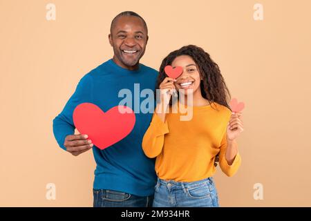 L'amour est dans l'air. Couple noir romantique avec coeur en papier rouge dans les mains posant sur fond de studio pêche Banque D'Images