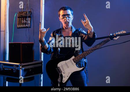 Un musicien fou qui chante du rock avec les doigts et criant à voix haute en studio, jouant de la guitare et chantant de la musique en métal lourd. Un artiste alternatif sympa qui interprète une chanson punk rock, en cuir. Banque D'Images