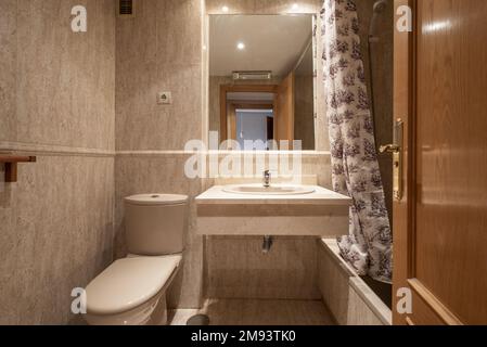 Salle de bains carrelée de marbre crème avec miroir intégré et toilettes en porcelaine et baignoire avec rideaux Banque D'Images