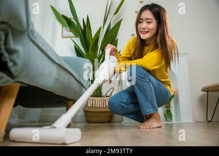 Bonne jeune femme asiatique avec aspirateur à accumulateur à la maison dans le salon Banque D'Images