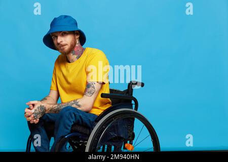 Un homme en fauteuil roulant a des problèmes avec le système musculo-squelettique regarde la caméra dans un t-shirt avec des tatouages sur ses bras assis sur un studio bleu Banque D'Images