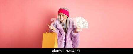 Une grand-mère riche et élégante qui fait du shopping avec des dollars, tient un sac en papier et sourit sans souci, se tenant sur un fond rose Banque D'Images