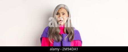 Gros plan d'une femme asiatique âgée choquée qui resplense, regardant avec un visage confus, regardant avec des cringe, se tenant sur un fond blanc Banque D'Images