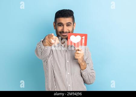 Évaluez-vous ma publication sur le réseau social ? Homme souriant blogueur tenant comme contre-signe et pointant le doigt vers l'appareil photo, portant une chemise rayée. Studio d'intérieur isolé sur fond bleu. Banque D'Images