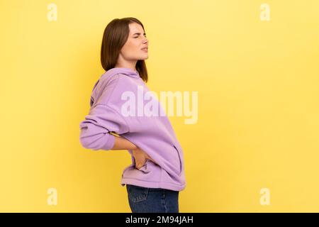 Portrait de profil de la femme anxieuse frustrée malsaine tenant les mains sur le bas du dos, une blessure aux reins, mal de dos, portant le capuchon violet. Studio d'intérieur isolé sur fond jaune. Banque D'Images