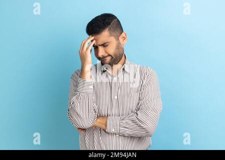 Homme d'affaires avec la barbe faisant le geste de visage gardant la main sur la tête, se blâmant pour la mauvaise mémoire, erreur impardonnable, portant une chemise rayée. Studio d'intérieur isolé sur fond bleu. Banque D'Images