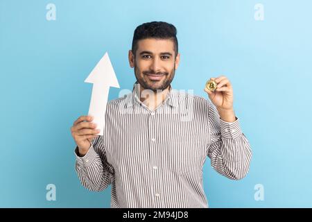 Portrait d'un jeune homme d'affaires adulte attrayant montrant une augmentation de croissance du bitcoin, exprimant des émotions positives, portant une chemise rayée. Studio d'intérieur isolé sur fond bleu. Banque D'Images