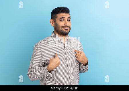 Portrait d'un homme d'affaires barbu se pointant et regardant égoïste haughty égoïste, se sentant fier de sa propre réalisation, portant une chemise à rayures. Studio d'intérieur isolé sur fond bleu. Banque D'Images