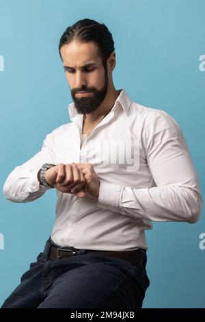 Portrait d'un homme sérieux aux cheveux sombres et attrayant avec une barbe portant une chemise blanche assis et regardant sa montre-bracelet, étant harry à un événement important. Studio d'intérieur isolé sur fond bleu. Banque D'Images