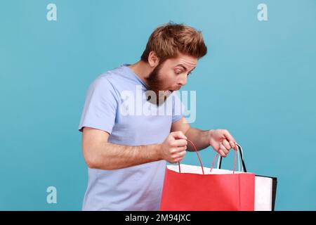 Portrait d'un bel homme barbu debout, tenant des sacs de shopping rouges et blancs et regardant à l'intérieur avec l'expression du visage choqué. Studio d'intérieur isolé sur fond bleu. Banque D'Images