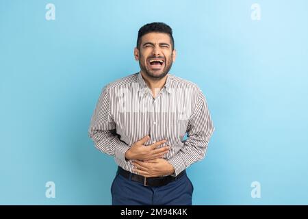 Portrait de drôle beau barbu jeune homme d'affaires debout avec les yeux fermés et rire, tenant son ventre, portant une chemise rayée. Studio d'intérieur isolé sur fond bleu. Banque D'Images