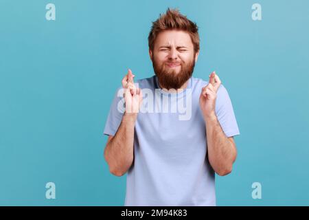 Portrait de l'homme barbu croisant les doigts pour la chance, faisant un souhait, rêvant de l'intérieur, rituel, se sentant plein d'espoir en attendant le miracle. Studio d'intérieur isolé sur fond bleu. Banque D'Images