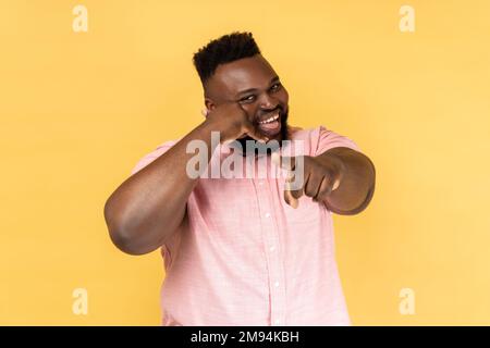 Portrait d'un homme souriant portant une chemise rose tenant les doigts près de l'oreille montrant le geste du téléphone regardant et pointant vers l'appareil photo, en attendant votre appel. Studio d'intérieur isolé sur fond jaune. Banque D'Images