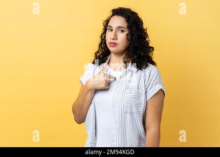 Portrait d'une femme sûre d'elle-même sérieuse avec des cheveux ondulés foncés pointant vers elle-même, se sentant fière et auto-important, ayant le grand ego. Studio d'intérieur isolé sur fond jaune. Banque D'Images