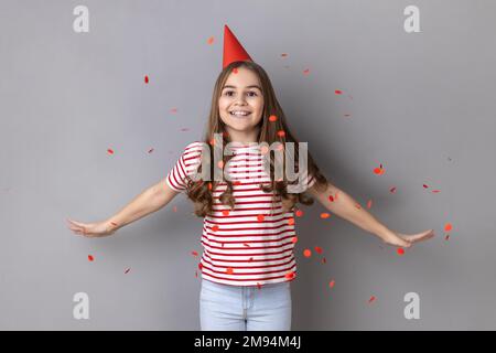 Portrait de charmante petite fille extrêmement heureuse portant un T-shirt rayé debout avec bras étalé sous des confettis tombant, célébrant l'anniversaire. Prise de vue en studio isolée sur fond gris. Banque D'Images