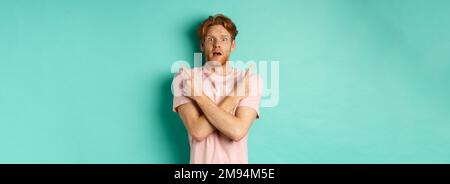 Homme nerveux avec des cheveux rouges épris indécise, bras croisés et pointant sur le côté, montrant deux variantes avec le visage perplexe, debout dans le t-shirt sur la menthe Banque D'Images