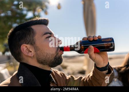 Vue latérale d'un jeune homme barbu de la bière d'une bouteille de verre à la lumière du soleil tout en passant le week-end sur la plage Banque D'Images