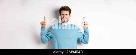 Déçu le modèle masculin frognant et se portant, pointant les doigts vers le haut avec une émotion négative, n'aime pas l'offre promo, se tenant sur fond blanc Banque D'Images