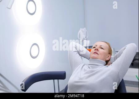 femme adulte dans la clinique dentaire attendant le médecin elle a mis ses mains derrière sa tête fauteuil bureau de repos belle lumière éclairage de bureau bonne réparation dans la clinique dentaire dernière technologie Banque D'Images