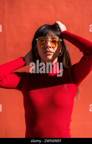 Portrait d'une fille brune avec ses mains sur son visage portant une robe orange et des lunettes de soleil au printemps, profitant d'une matinée ensoleillée. Plutôt caucasien Banque D'Images