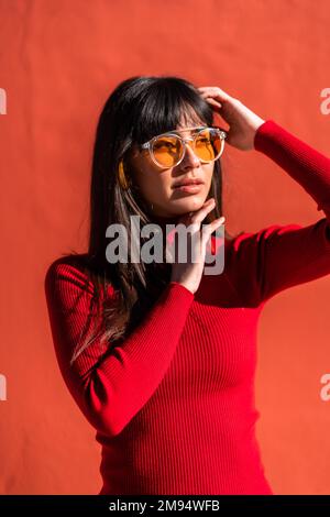 Portrait d'une fille brune avec ses mains sur son visage portant une robe orange et des lunettes de soleil au printemps, profitant d'une matinée ensoleillée. Plutôt caucasien Banque D'Images