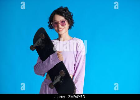 Portrait d'une adolescente souriante tenant un skateboard, isolée sur fond bleu Banque D'Images