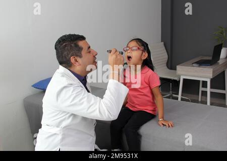 Médecin pédiatre professionnel et patient jeune fille à poil brun Latino de 4 ans sont en consultation pour vérifier leurs yeux, leur bouche et leur gorge Banque D'Images