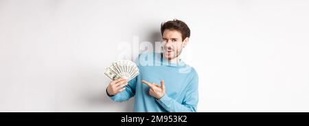 Beau gars riche qui se vante à la caméra, montrant des revenus, pointant le doigt sur l'argent gagné et souriant, debout sur fond blanc Banque D'Images