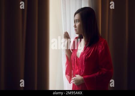 Ouverture de la femme heureuse dans une chemise de nuit rouge ouvrant les rideaux de fenêtre. Banque D'Images