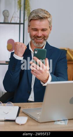 Heureux étonné homme d'affaires mature travaillant sur smartphone choqué par la victoire soudaine du jeu gagnant de loterie jackemen bonnes nouvelles, la croissance de carrière. Homme mi-âgé aux cheveux gris pigiste au bureau à domicile Banque D'Images