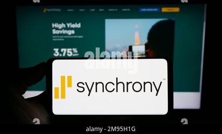 Personne tenant un téléphone portable avec le logo de la société de services financiers synchrony Bank à l'écran en face de la page Web d'affaires. Mise au point sur l'affichage du téléphone. Banque D'Images