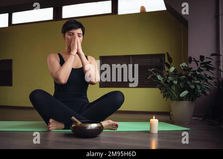Une femme d'âge moyen, pratiquant le yoga, effectue une méditation assise sur un tapis dans une pose de lotus avec un namaste, s'entraîne dans une salle avec Pranayama Banque D'Images