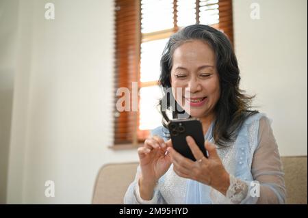 Charmante et heureuse femme asiatique de 60s ans utilisant son smartphone, discutant avec ses amis tout en se relaxant dans son salon. Le style de vie de la retraite concep Banque D'Images