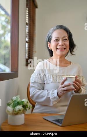 Portrait, charmante femme asiatique 60s à la retraite tenant une tasse de thé, souriant et regardant l'appareil photo tout en se relaxant dans le café. Banque D'Images