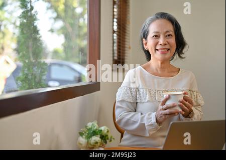 Charmante et heureuse 60s femme asiatique à la retraite tenant une tasse de thé, souriant et regardant la caméra tout en se relaxant dans le café. Banque D'Images