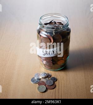 Un vieux pot en verre rempli d'économies de beaucoup de pièces sur une table en bois. Certaines pièces sont sur la table Banque D'Images