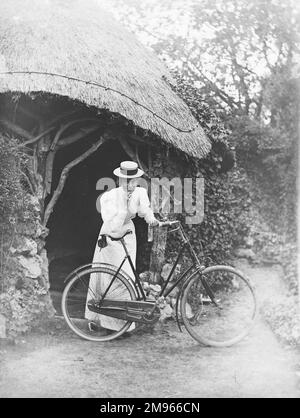 Une dame en robe d'été blanche longue et de boater se penche sur son vélo droit à l'entrée d'une maison d'été ou d'une grotte dans le domaine d'une grande maison de campagne Banque D'Images