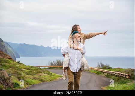 Un jeune couple marche sur une route entourée par la nature, avec l'homme portant la femme sur son dos Banque D'Images