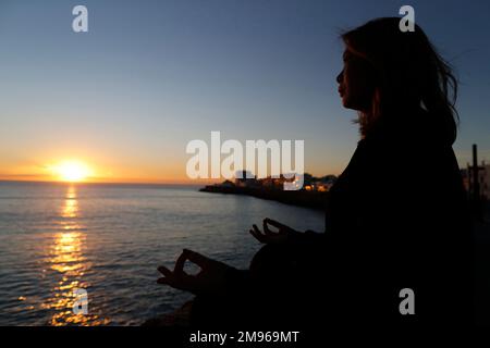 Femme pratiquant la méditation de yoga par la mer au coucher du soleil comme concept de silence et de détente. Banque D'Images